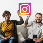 Duas pessoas sorrindo sentadas em um sofá com fones de ouvido e smartphones, segurando um ícone do Instagram.