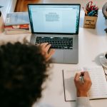 Pessoa escrevendo conteúdo de marketing no laptop em mesa de trabalho com caderno e café